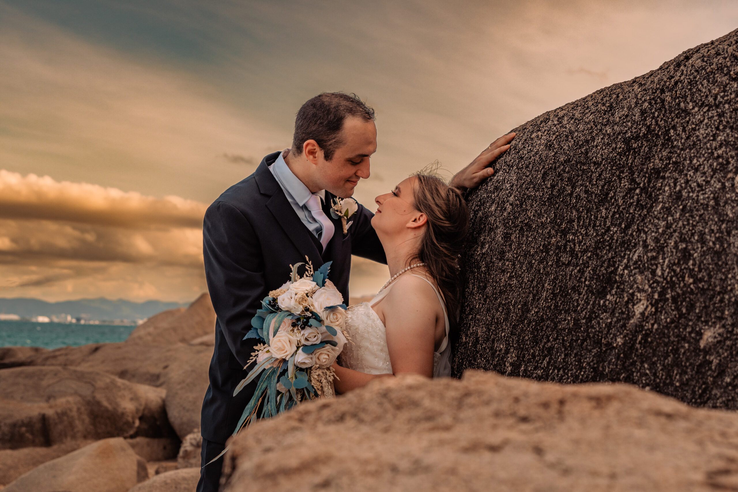bride and groom staring at each other while leaning against a rock wall at sunset on their wedding day