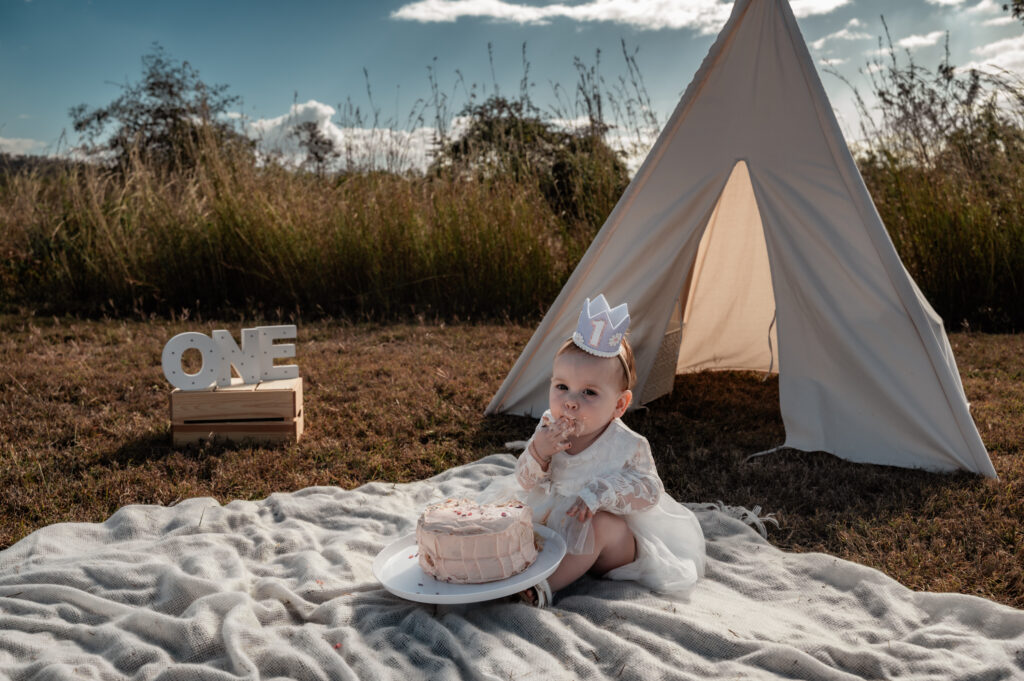 baby girls celebrating her 1st birthday by having a cake smash in a paddock with a teepee behind her - milestone photography by Jamie Simmons
