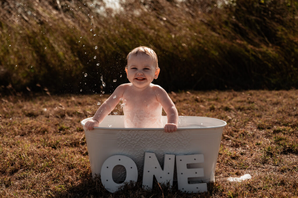 baby girl splashing water in her bubble bath out in a paddock - milestone photography by Jamie Simmons