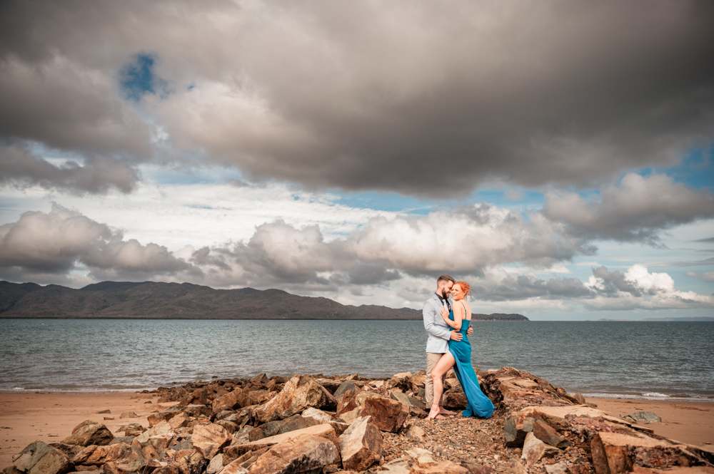 man whispers something in fiancé's ear as she laughs while they embrace on a rock wall on a townsville beach - engagement photography by Jamie Simmons