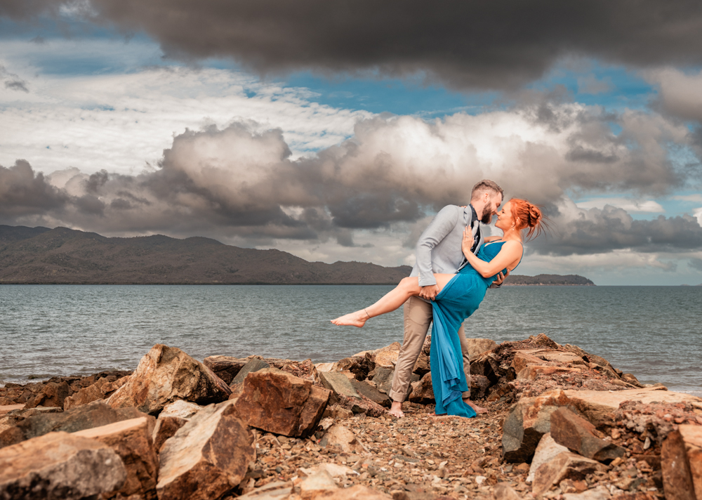 man and woman standing on a rock wall at a townsville beach as the man holds fiancé's leg and dips her - engagement photography by Jamie Simmons