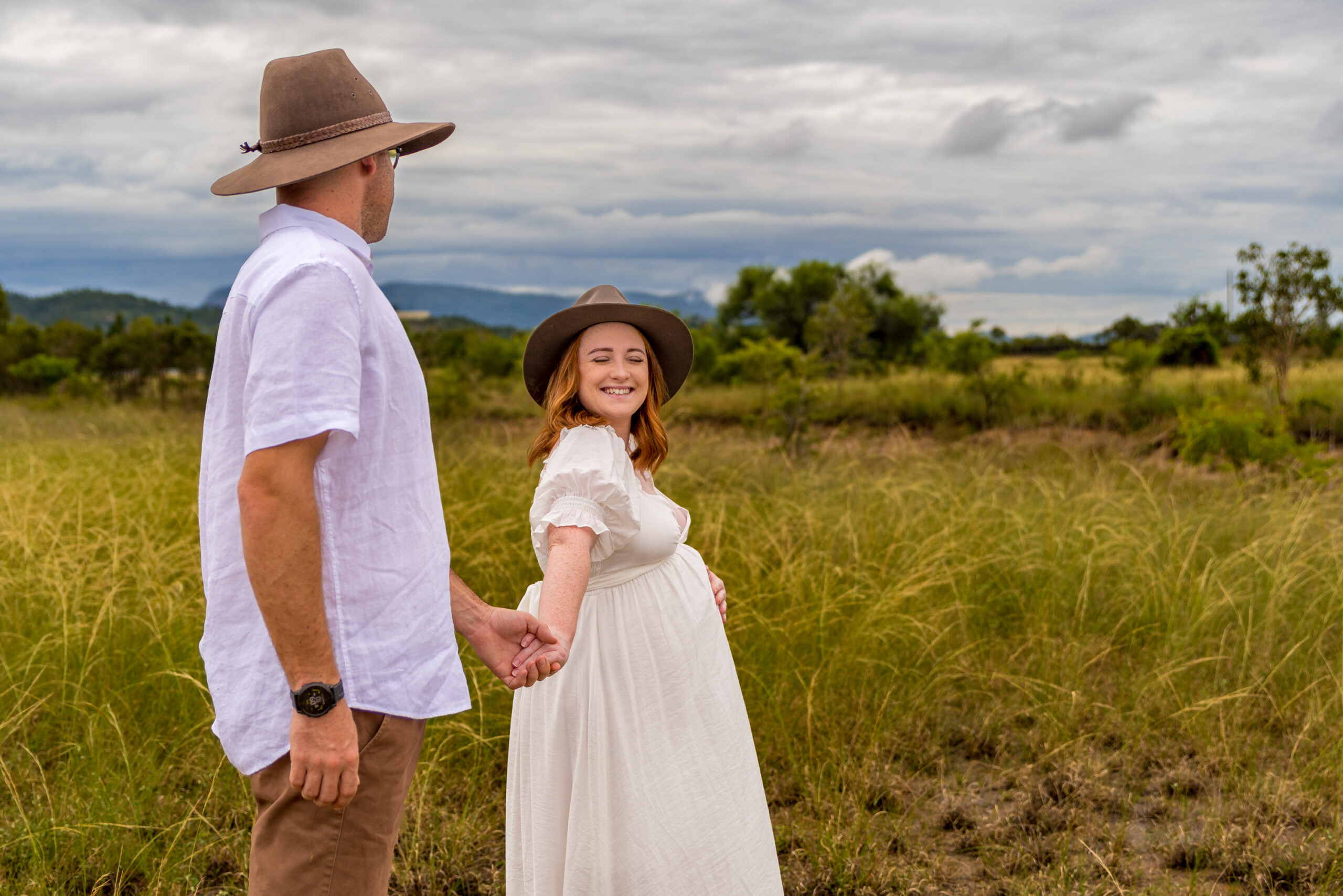 pregnant woman in white maternity gown holding her baby belly and leading her partner through a townsville paddock while looking back at her partner smiling - maternity photography by Jamie Simmons