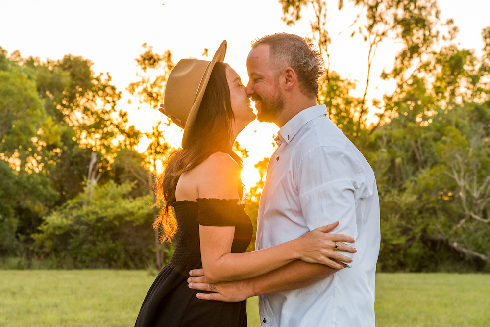 woman in black dress and man in white shirt embracing and leaning in for a kiss as the setting sun shines through - engagement photography by Jamie Simmons