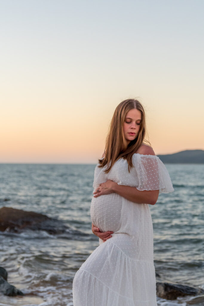 pregnant woman in white maternity gown embracing baby belly while at the beach at sunset - maternity photography by Jamie Simmons