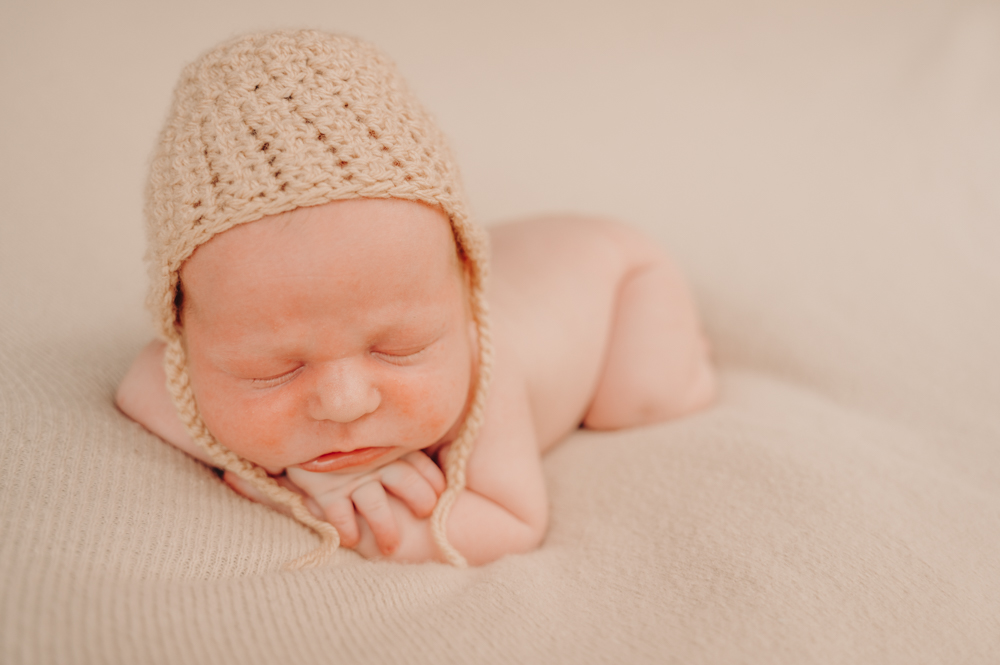 nude newborn baby wearing a crocheted beanie lying on cream blanket and sleeping on his hands - newborn baby photography by Jamie Simmons