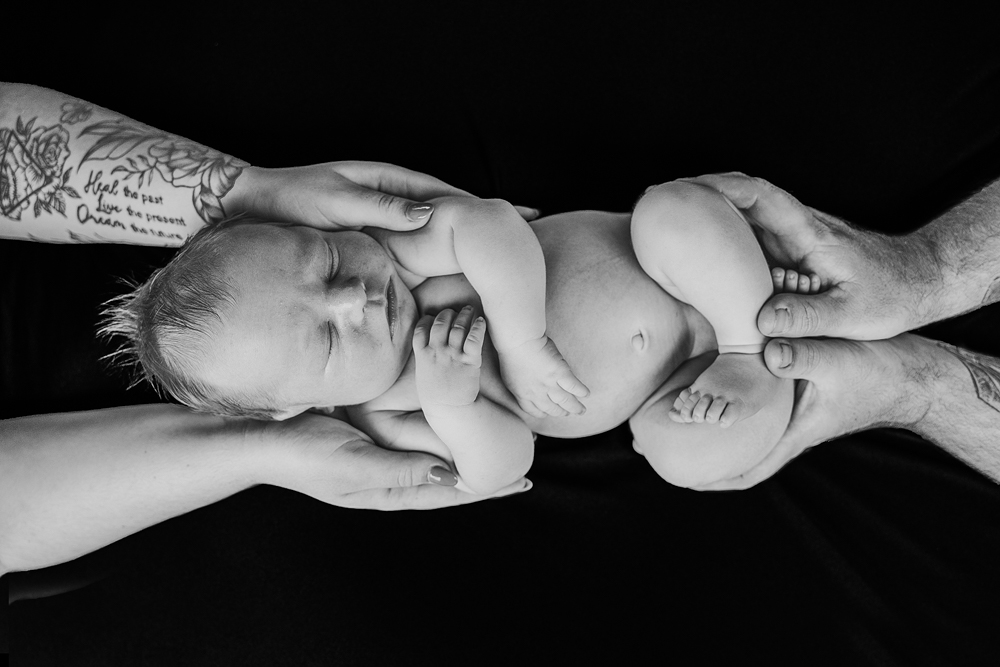 monochrome shot of mum and dad holding newborn between them over a black background - newborn photography by Jamie Simmons