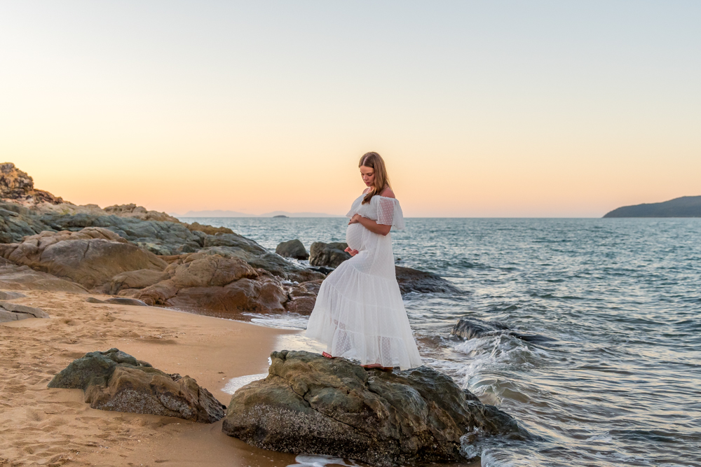 woman in white maternity gown standing on rocks on the beach at sunset cradling baby belly - maternity photography by Jamie Simmons
