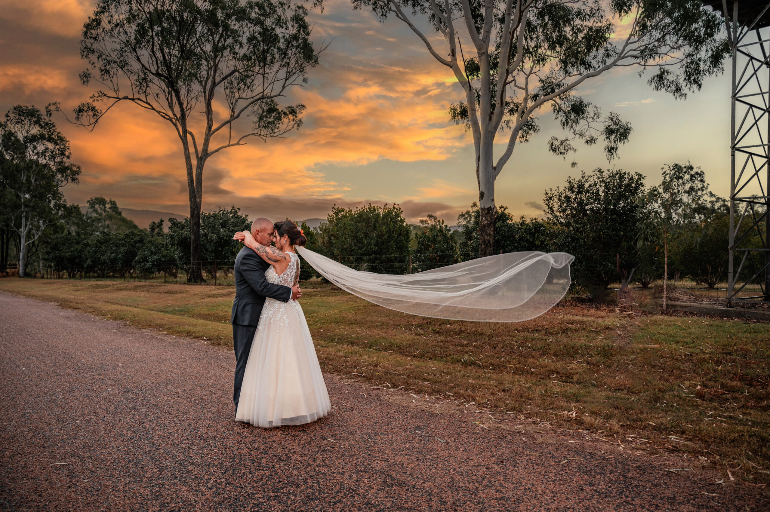 husband and wife embracing on country road at sunset with brides veil blowing in wind