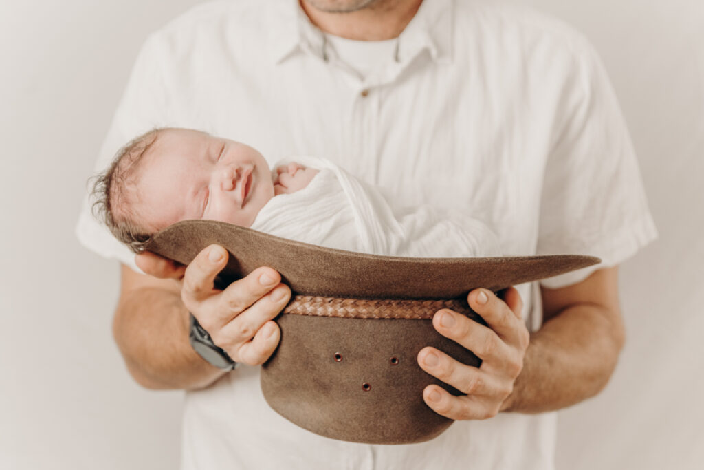 newborn baby wrapped and posed inside cowboy hat being held by dad - newborn photography by Jamie Simmons