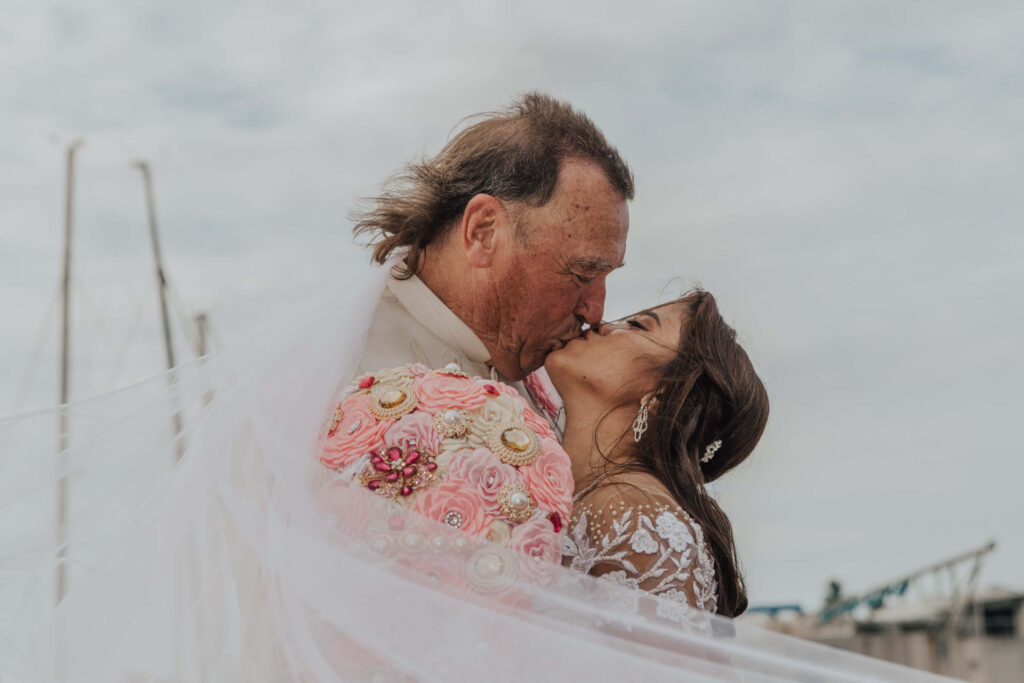 A bride and groom share a romantic kiss on a picturesque dock, surrounded by serene water and a beautiful backdrop. - Photo by Jamie Simmons