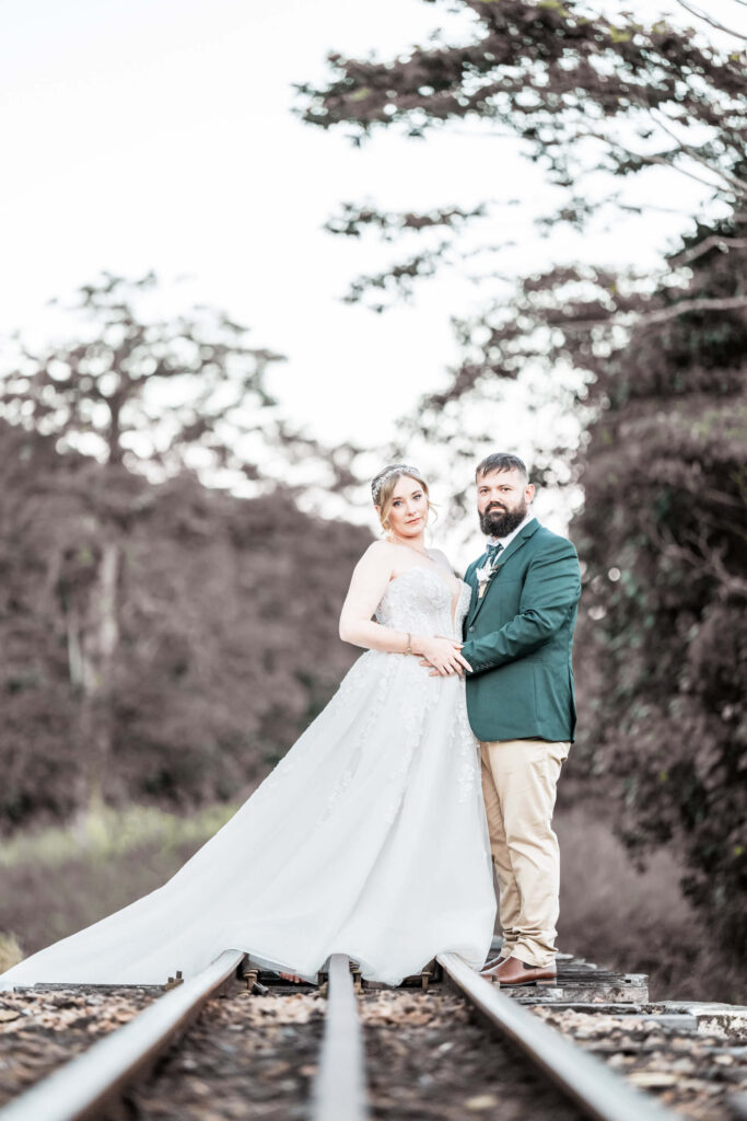 The bride and groom stand on rustic train tracks, enveloped by trees, embodying romance and the journey of marriage. - by Jamie Simmons