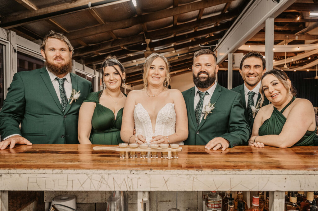 A joyful wedding party gathers at the bar, smiling and posing for a memorable group photo. - by Jamie Simmons