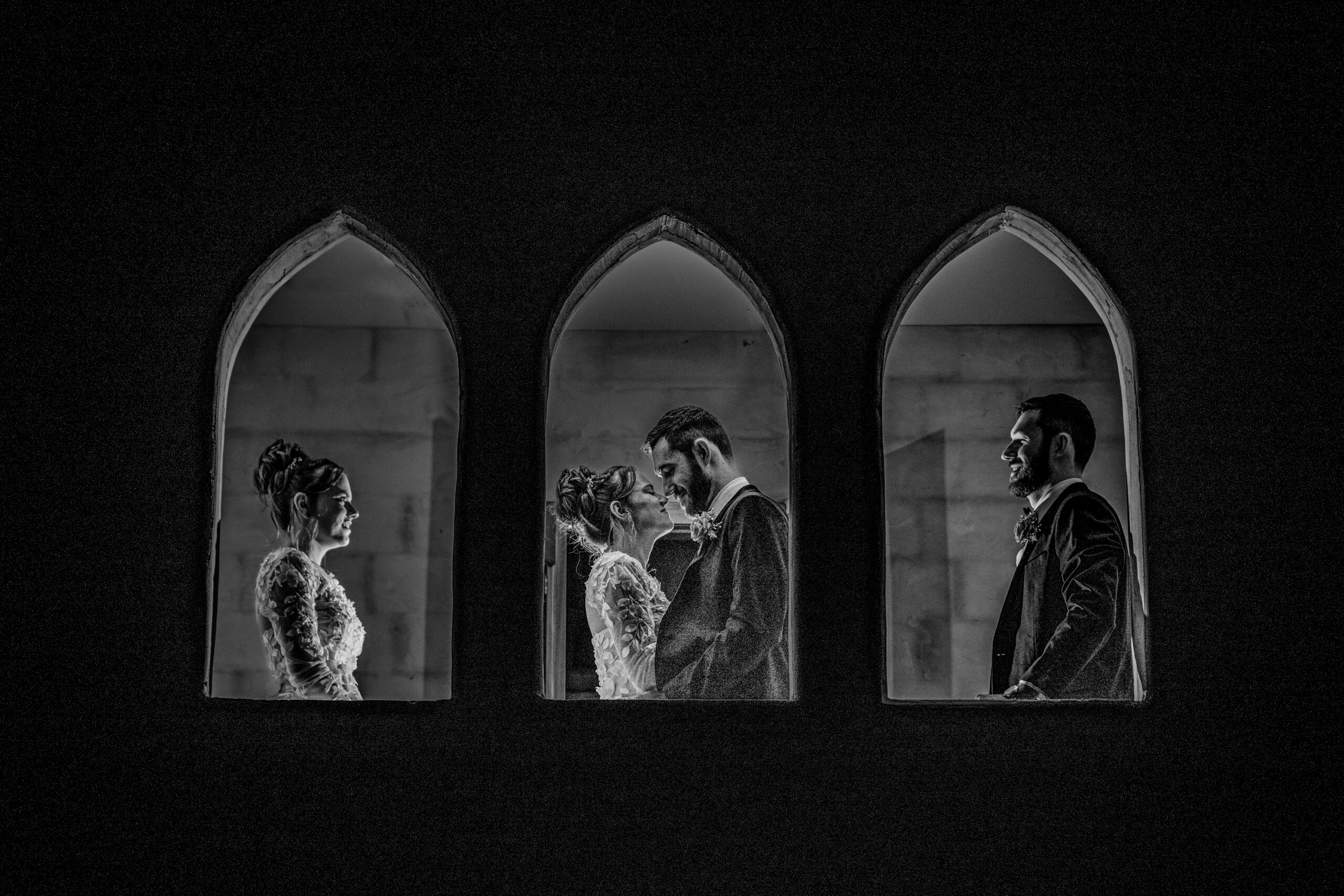 A black and white photo by a Townsville wedding photographer shows a bride and groom in formal attire, framed by three arched windows, standing face to face and holding hands.