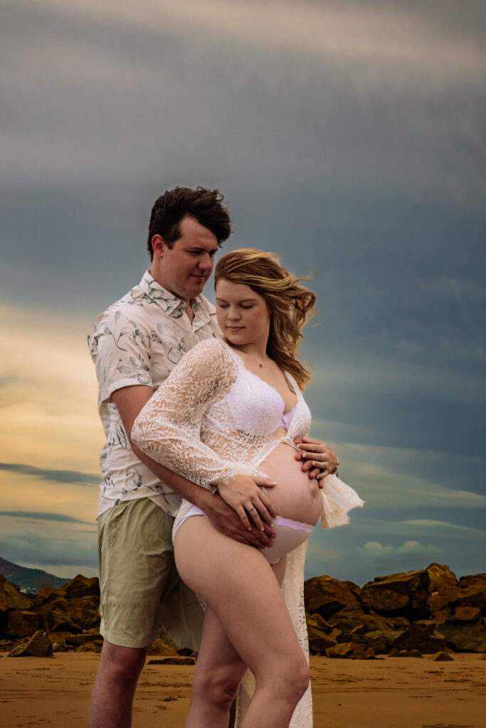 A couple stands on a rocky beach at sunset. The woman, pregnant and in a white lace outfit, is embraced by her partner as he holds her belly—an intimate scene captured by Townsville Motherhood photographer Simmons Memorable Moments.