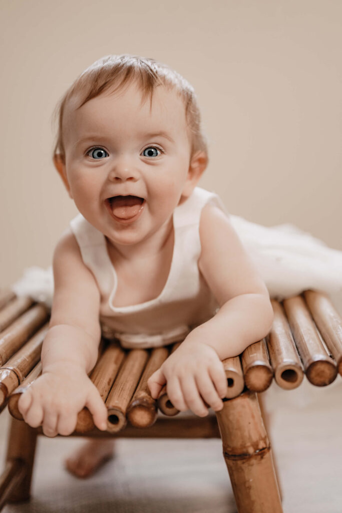 Smiling baby with bright blue eyes lying on a bamboo bench, wearing a soft white outfit against a neutral beige background; joyful infant portrait with open mouth expression, natural light studio photography, close-up baby milestone session, warm earthy tones, Townsville baby photographer Simmons Memorable Moments, candid childhood moment, professional newborn and baby photography.