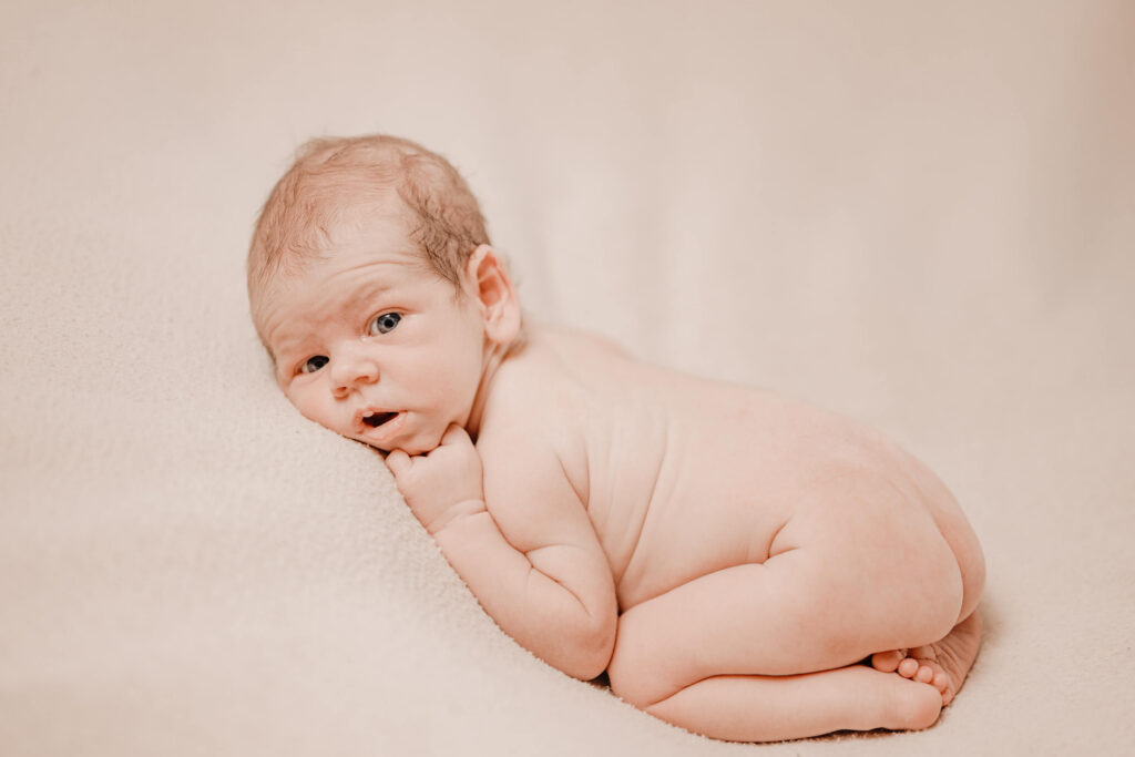 A newborn baby lies on a soft beige blanket, resting on their stomach with their head turned to the side and eyes open—captured beautifully by Townsville photographer Simmons Memorable Moments